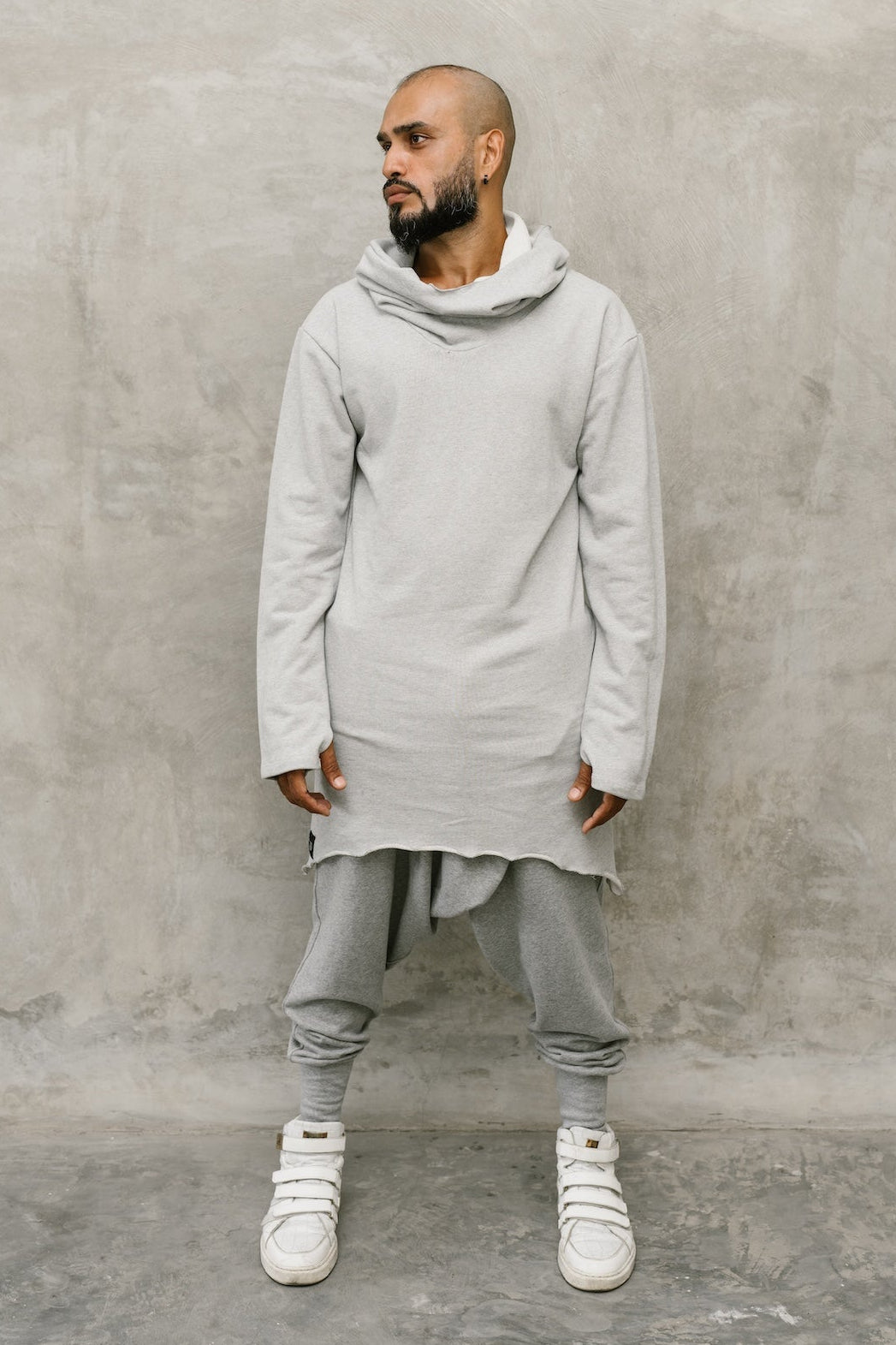 A man wearing a light gray oversized long-sleeve hoodie, matching gray jogger pants, and white sneakers, standing against a textured gray concrete wall.