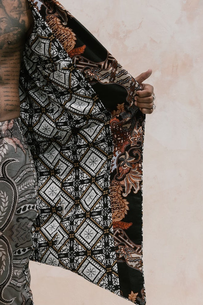 A shirtless man with a bun stands by a pool, holding open a large patterned textile draped like a cape over his shoulders. He is wearing matching patterned pants and black boots, against a pale stucco wall with white steps and ceramic pots.