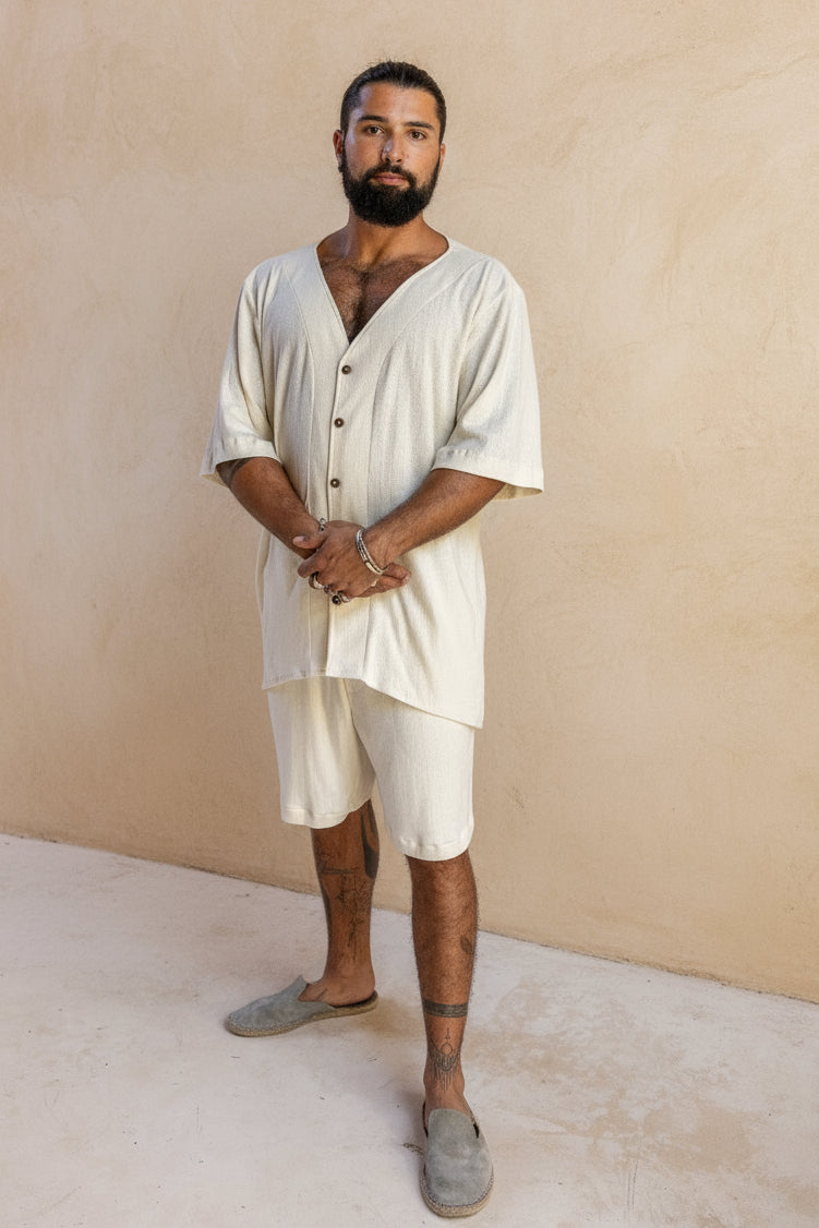 A person standing in a traditional hut setting, wearing an ivory white sleeveless cotton shirt with unique wooden buttons and medium long sleeves, paired with shorts.