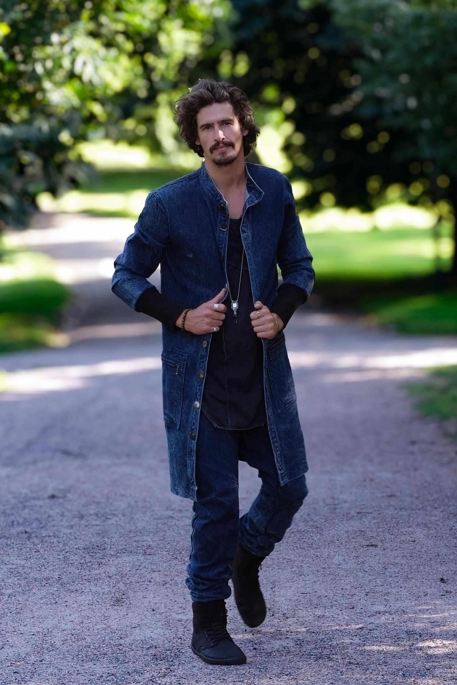 Man wearing a denim jacket and jeans standing on a gravel path with greenery in the background