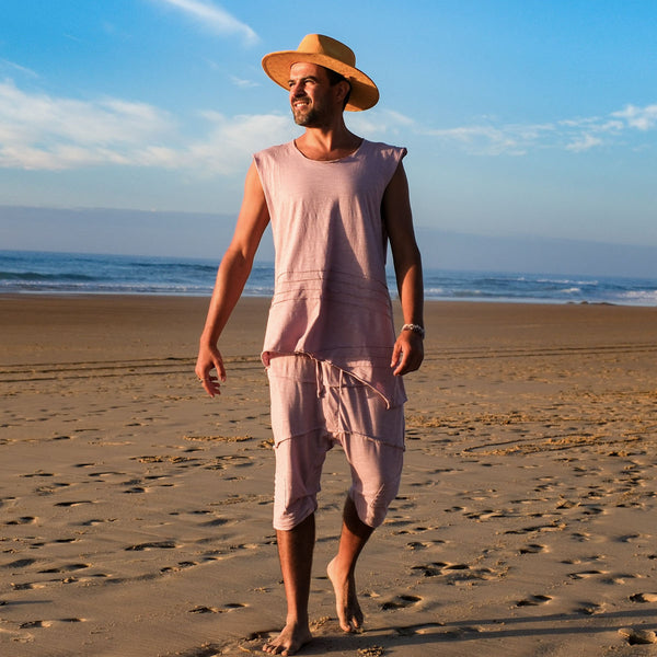 Man walking on a beach wearing a sleeveless pink top and beige hat.