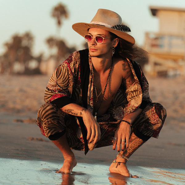 Person in bohemian attire squatting on a beach with palm trees in the background