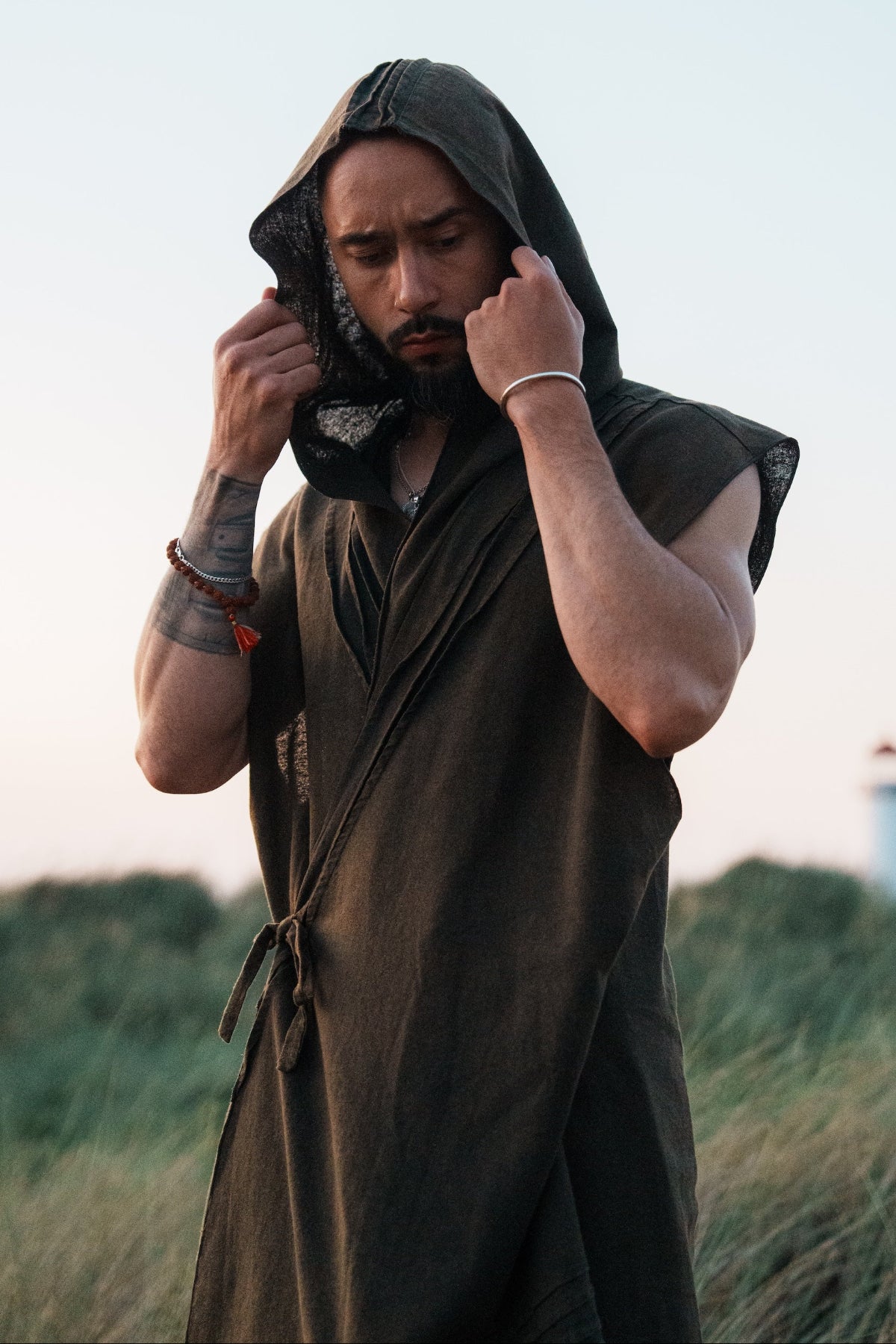 A man wearing a dark olive green hooded robe stands in a grassy outdoor setting at dusk, holding the hood up around his face. A lighthouse is faintly visible in the background under a pale sky.