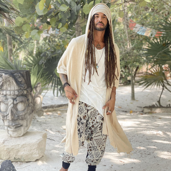 Man with long dreadlocks wearing a beige robe and patterned pants in a tropical setting.
