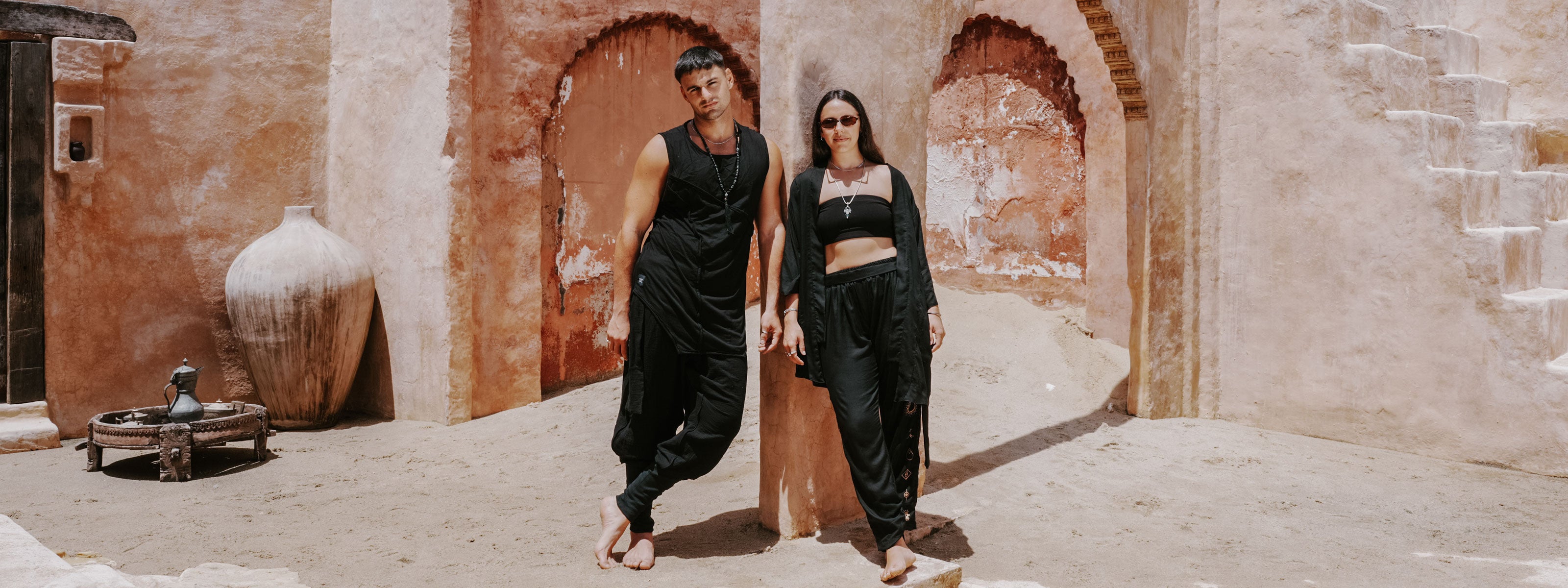 A man and a woman wearing matching black outfits pose together in front of rustic desert-style adobe walls with arches.