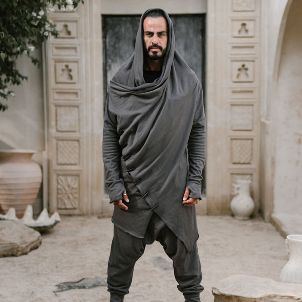 Man wearing a long, dark gray hooded garment standing in an outdoor setting with architectural elements.