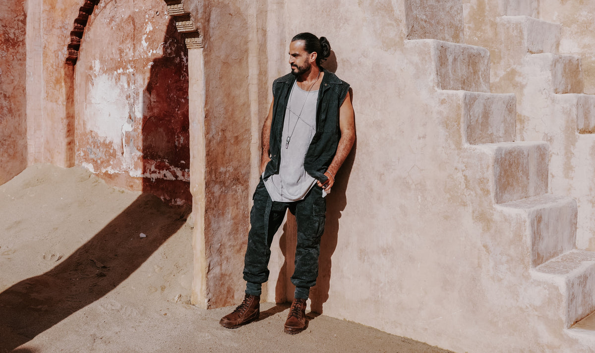 A man stands outdoors against a sunlit beige plaster wall and stone stairs, wearing a sleeveless dark vest over a light gray T-shirt, dark pants, and brown boots.