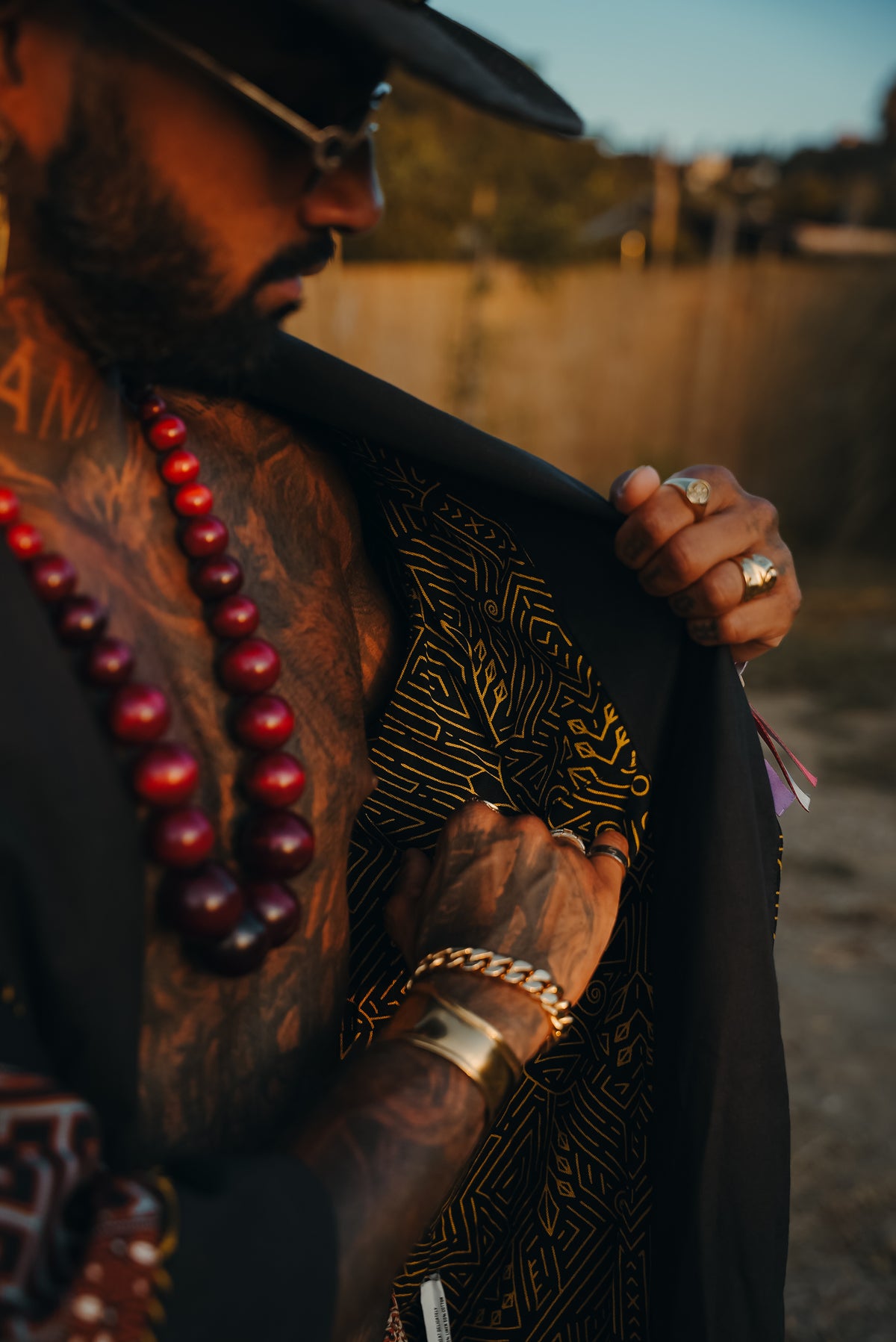 A close-up lifestyle shot of a tattooed man wearing a black fedora, gold chain bracelets, and red beaded necklaces, holding open a black jacket with a gold patterned lining.